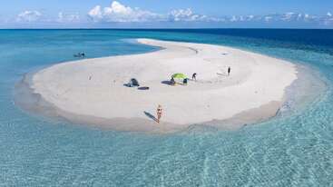 Eine kleine, abgelegene Sandbank, umgeben von klarem, blauem Meer. Mehrere Menschen entspannen sich unter Sonnenschirmen und Zelten und genießen die ruhige, unberührte Schönheit dieses abgelegenen tropischen Paradieses.