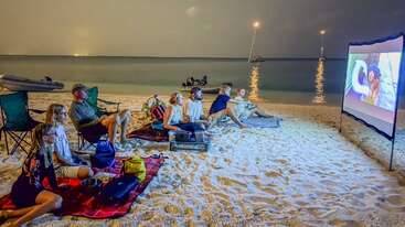 Eine Gruppe von Leuten sitzt nachts am Sandstrand und schaut sich einen Film an, der auf eine Leinwand projiziert wird, während im Hintergrund das Meer und Boote zu sehen sind.