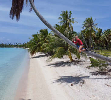 Eine Frau in einem roten Shirt und blauen Shorts klettert auf eine Palme an einem tropischen Strand mit weißem Sand und türkisfarbenem Wasser unter blauem Himmel.