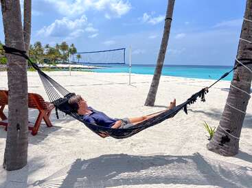 Jemand entspannt sich in einer Hängematte, die zwischen Palmen an einem sonnigen Strand gespannt ist. Weißer Sand, türkisfarbenes Wasser, ein Volleyballnetz und ein strahlend blauer Himmel bilden ein Paradies.