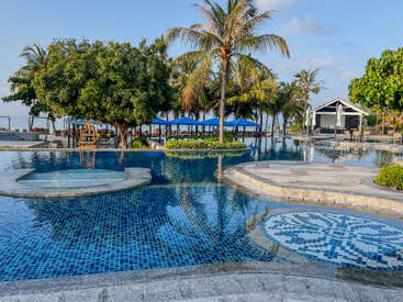 Une scène paisible autour de la piscine d'un complexe hôtelier, avec des palmiers, des carreaux de mosaïque bleus, des chaises longues, des parasols et une eau limpide reflétant le ciel serein, invitant à la détente et au farniente.