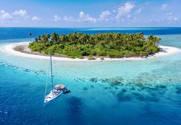Une petite île tropicale aux palmiers luxuriants est entourée d'eaux turquoise. Un voilier blanc est ancré tout près, sous un ciel bleu parsemé de nuages.