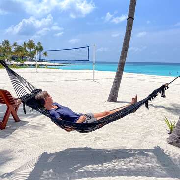 Jemand entspannt sich in einer Hängematte, die zwischen Palmen an einem sonnigen Strand gespannt ist. Weißer Sand, türkisfarbenes Wasser, ein Volleyballnetz und ein strahlend blauer Himmel bilden ein Paradies.
