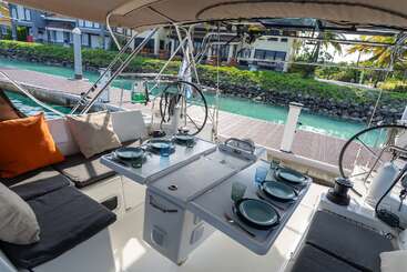 Un coin repas chaleureux sur un yacht, dressé avec assiettes et verres, donne sur une eau turquoise, un quai et de charmantes maisons entourées d'une végétation luxuriante et de palmiers.
