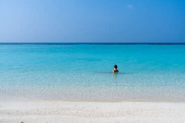 Une personne se détend dans une mer turquoise, limpide et peu profonde, près d'une plage de sable blanc, sous un ciel bleu serein, profitant d'une solitude paisible et de la beauté de la nature.