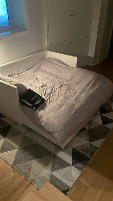 A neatly made bed with light bedding sits against a wall, next to a window. A geometric rug covers the wooden floor. A bag lies on the bed.