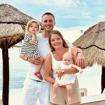 A smiling family of four poses by the seaside, under thatched umbrellas. The parents hold their young daughters, enjoying a bright, relaxing day together on vacation.