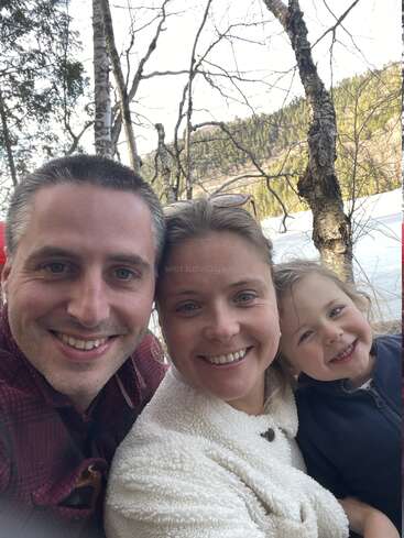 Una familia de tres miembros, con una sonrisa en el rostro, posa para una selfie en primer plano al aire libre, con árboles, un lago y colinas verdes al fondo. Todos parecen felices juntos.