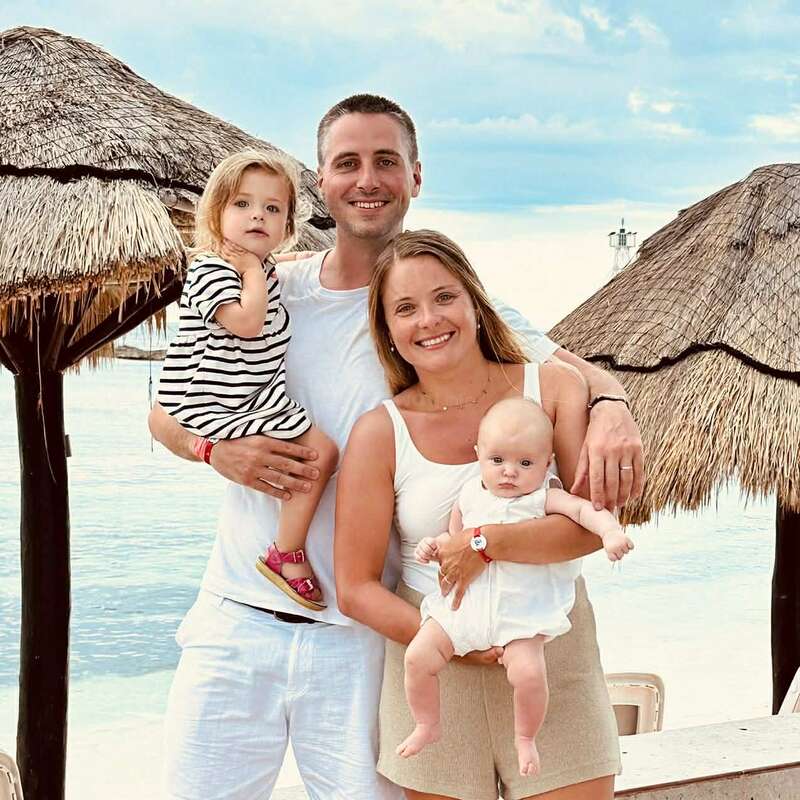 A smiling family of four poses by the seaside, under thatched umbrellas. The parents hold their young daughters, enjoying a bright, relaxing day together on vacation.