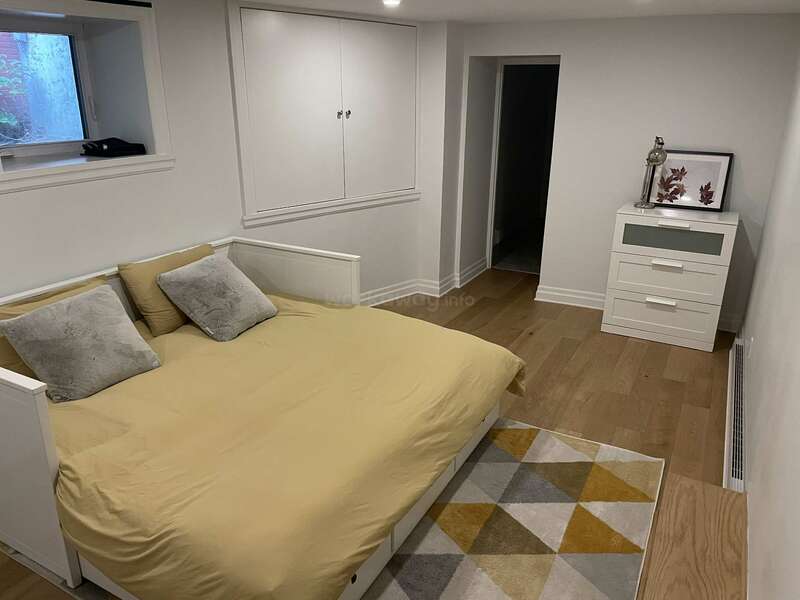 Este acogedor dormitorio cuenta con una cama con ropa de cama beige y almohadas grises, una alfombra geométrica, suelos de madera, una ventana, cajones blancos y sencillos detalles decorativos.