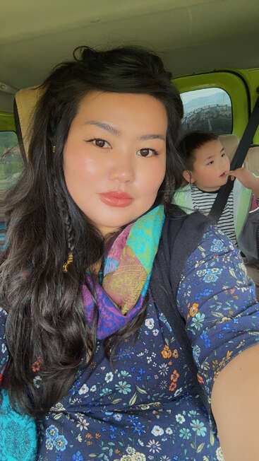 Una mujer de pelo largo y oscuro y vestido de flores se hace un selfi dentro de un coche. Detrás de ella hay un niño pequeño, vestido con ropa a rayas, que mira hacia fuera pensativo.