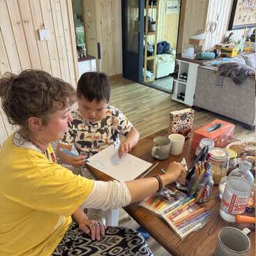 A woman and young boy are drawing together at a wooden table covered with colorful pens, mugs, snacks, and tissues in a cozy, organized home.