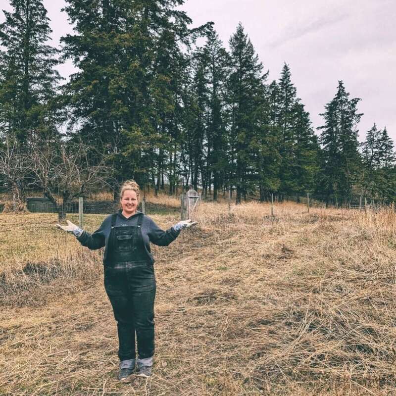 Une personne vêtue d'une salopette et de gants se tient debout, souriante, dans un champ herbeux, entourée d'herbe sèche et de grands arbres à feuilles persistantes, sous un ciel couvert, les bras tendus.