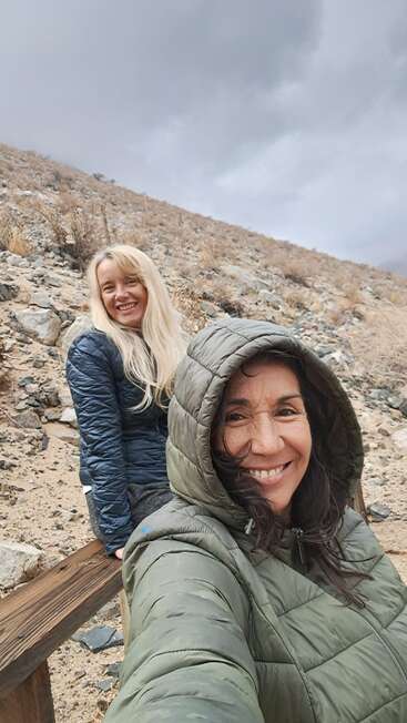 Two women enjoy a hike on a rocky hillside, smiling and bundled up in jackets. The sky is cloudy, and the landscape appears dry and rugged.