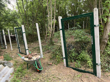 Uma cerca de arame recém-instalada, com portões de metal, fica em uma área arborizada. Um carrinho de mão cheio de pedras e madeira está parado ali perto, no chão de terra.