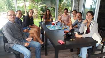 A imagem mostra um grupo de pessoas reunidas em torno de uma mesa, provavelmente jogando um jogo de tabuleiro, com uma mesa e cadeiras visíveis ao fundo, transmitindo uma sensação de interação social e lazer.