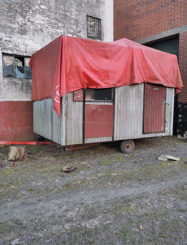 A small, weathered wooden shed with wheels is covered by a red tarp. It stands outside against a stained, old wall beside broken and dirty windows.