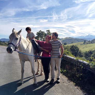 Un niño pequeño va sentado en un caballo blanco, guiado por dos adultos. Están al aire libre en un día soleado, con verdes colinas y un cielo azul de fondo.