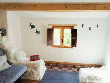 This cozy room has white textured walls, wooden beams, a window with shutters, built-in seating, turtle wall decor, patterned tile floor, and a blue couch.