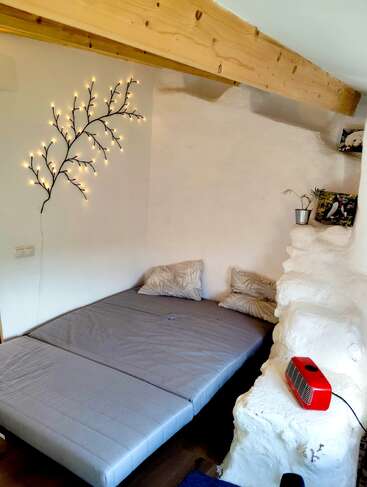 A cozy rustic bedroom features a low bed with gray bedding, decorative branch string lights, wooden beams, white textured walls, plants, photos, and a red heater.