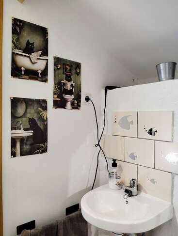 This image shows a bathroom with a small sink, fish-decorated tiles, cat-themed wall art, a towel, liquid soap, and a metal bucket on a ledge.
