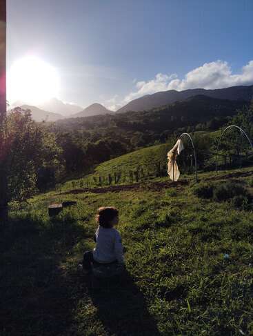 A young child sits on grass in a sunlit rural landscape, surrounded by green hills, trees, distant mountains, and blue sky with fluffy clouds. Peaceful moment.