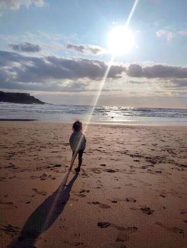 A child stands alone on a sandy beach, gazing at the sparkling ocean. The bright sun casts a long dramatic shadow, creating a peaceful atmosphere.
