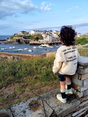A child stands on a stone wall, overlooking a scenic harbor with boats, grassy fields, and charming houses under a blue sky, wearing a statement sweater.