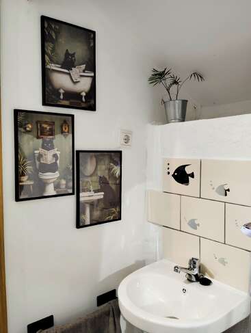 Cette salle de bains présente un art mural excentrique sur le thème des chats, des carreaux décorés de poissons au-dessus d'un lavabo blanc, une serviette grise, une petite plante en pot et un design épuré et minimaliste.