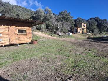 Une cabane rustique en bois se trouve sur un terrain herbeux, avec un chemin de terre menant à une autre maison en bois à deux étages entourée d'arbres et de collines sous un ciel bleu.