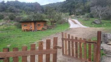 Uma cabana rústica de madeira fica em um campo gramado, cercada por árvores e colinas. Um portão de madeira aberto leva a uma estrada de terra e a uma paisagem tranquila.
