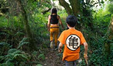 Um menino com uma camisa laranja com um símbolo japonês segue uma mulher por uma floresta verdejante, explorando a natureza juntos num dia ensolarado.
