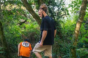 Um cara com dreadlocks e uma criança estão explorando uma floresta verdejante e exuberante. A criança usa uma camisa laranja e se abaixa com curiosidade entre as árvores e a folhagem.