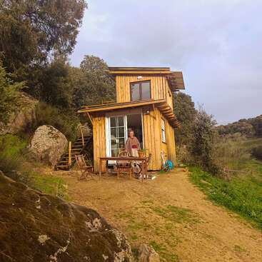 Uma pequena cabana de madeira fica em uma colina cercada por árvores. Uma pessoa está sorrindo do lado de fora, perto de uma mesa e cadeiras, curtindo o ambiente natural e tranquilo.