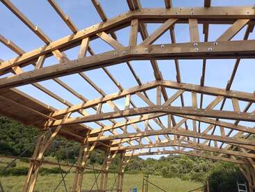 This image shows a wooden roof frame structure under construction, set outdoors with trees and grass in the background and a clear, blue sky above.