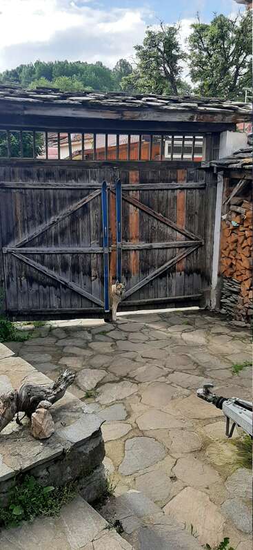 A imagem mostra uma cena rústica com um portão de madeira, um cachorro e uma pilha de lenha, em um fundo de pedra com uma cerca de madeira desgastada pelo tempo.