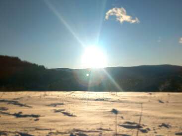 A imagem mostra um sol brilhante nascendo ou se pondo sobre um campo nevado, projetando sombras longas, com colinas e um céu azul claro ao fundo.