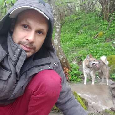 Un hombre vestido con una chaqueta con capucha y pantalones rojos está agachado en un frondoso bosque junto a un pequeño arroyo, con un perro detrás de él.