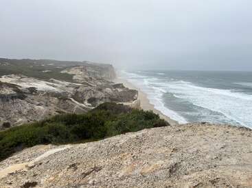 Felsige Klippen überblicken einen langen Sandstrand, an dem die Wellen brechen. Dichte Vegetation und ein nebliger, bedeckter Himmel schaffen eine ruhige, dramatische Küstenszene.