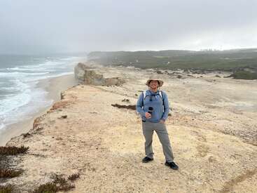 Ein Mann mit Hut und Outdoor-Kleidung steht lächelnd auf einer felsigen Küstenklippe. Unter ihm rauschen die Wellen des Meeres und der Himmel ist bedeckt und neblig.