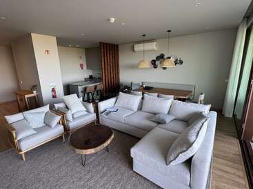 Dieses Bild zeigt ein modernes Wohnzimmer mit einem grauen Sektionssofa, zwei Sesseln, einem Couchtisch aus Holz, einer offenen Küche und einem Essbereich.