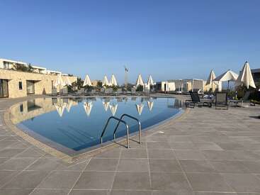 Ein ruhiger Swimmingpool, umgeben von leeren Liegestühlen und weißen Sonnenschirmen, in denen sich ein klarer blauer Himmel spiegelt. Moderne Steingebäude und Cabanas vervollständigen die heitere Szenerie.