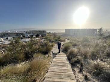 Ein Junge geht auf einem Holzweg durch grasbewachsene Sanddünen auf moderne Gebäude zu, während die helle Sonne in einen klaren Himmel scheint.
