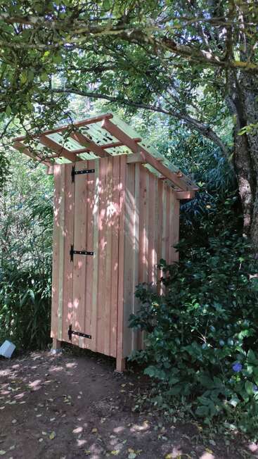 The image depicts a wooden outhouse situated in a wooded area, featuring a roof and door with black hinges, surrounded by lush greenery and trees.