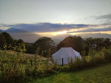 A white tent sits in a meadow surrounded by greenery and trees, with mountains in the background and a beautiful sunrise casting light through the clouds.