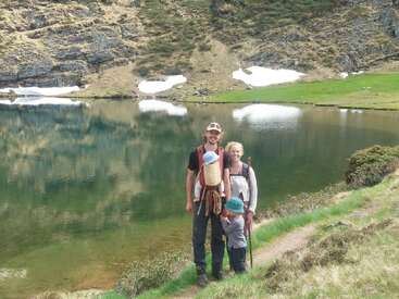 A smiling family of four, including two small children, enjoys a scenic hike near a tranquil mountain lake with snow patches and lush, grassy surroundings.