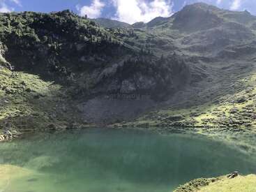 A serene, emerald lake reflects the lush, green mountains and scattered trees surrounding it under a bright blue sky with fluffy white clouds above. Peaceful scenery.