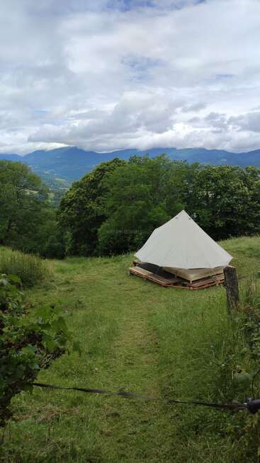 A white tent sits on wooden pallets in a green meadow, surrounded by trees with mountains in the distance under a partly cloudy sky.
