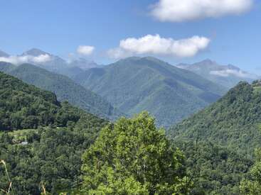 Lush green mountains, covered in dense forests, stretch into the distance. Soft clouds rest atop peaks under a clear blue sky, creating a peaceful, natural landscape.