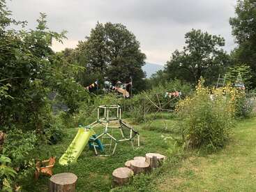A lush, green outdoor playground features a slide, climbing structures, wooden stepping stools, wild plants, and festive bunting. Large trees and cloudy sky in background.